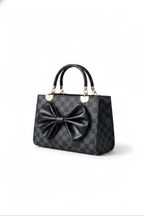 The Nocturne Bow Handbag is a black leather bag featuring a checkered design, gold hardware, two top handles, and a large black leather bow on the front, all showcased against a plain white background.